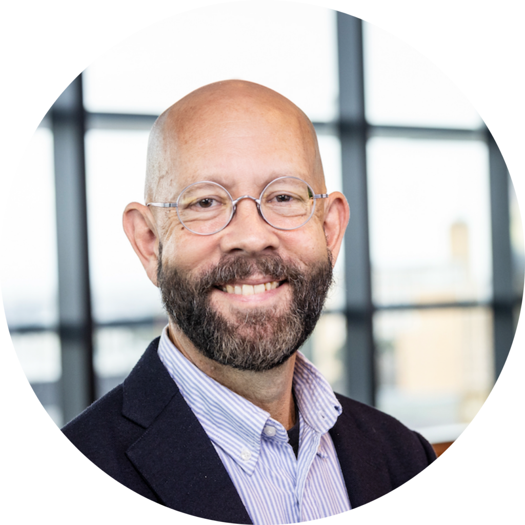 Portrait of Jeff Christiansen smiling, wearing round glasses, a dark beard and a navy jacket.